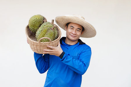 Handsome Asian man farmer wears hat, blue shirt, holds basket of durian fruits on shoulder. Concept,...