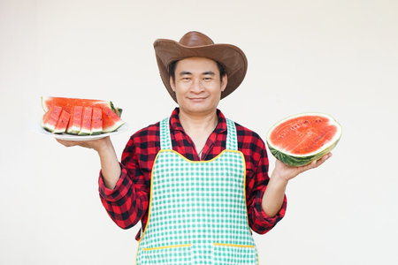 Handsome Asian man wears hat, apron, holds watermelon fruit to serve and present. Concept, Agriculture crops. Thai farmer, grow and sell watermelon fruits as economic crops.の写真素材