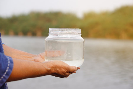 Closeup hands hold transparent jar that contain sample water from the lake. Concept, explore, inspect quality of water from natural source. Ecology survey. Environment conservationの写真素材