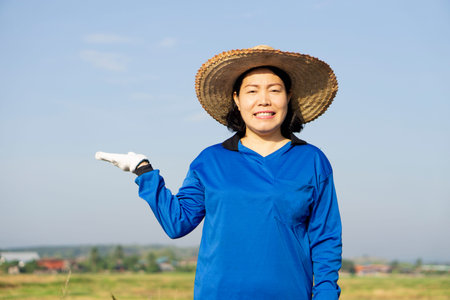 Portrait of Asian woman farmer wears hat and blue shirt, make gesture to present product for advertisement. Concept : Agriculture occupation. , Sky background.の写真素材