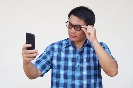 Asian man has problem with eyesight , wears eyeglasses, holds   smartphone. Concept, Eyesight problem. Optometry. Glasses with convex or concave lenses. Myopia.Eye diseaseの写真素材