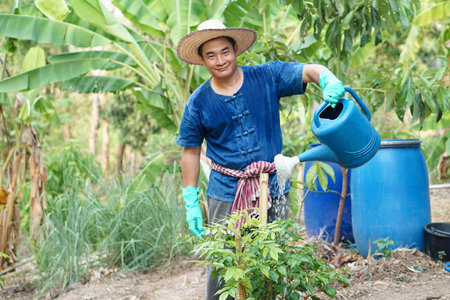 Asian man gardener holds blue watering can to water plants in garden. Concept, Gardening, taking care plant. Thai local agriculture lifestyle. Sufficiency economy. Work with natureの写真素材