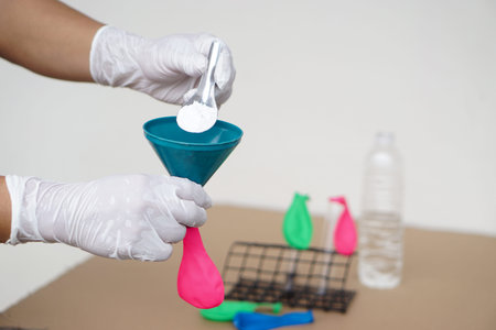 Closeup hands wear gloves holds funnel and flat pink balloon, put spoon of baking soda powder to...