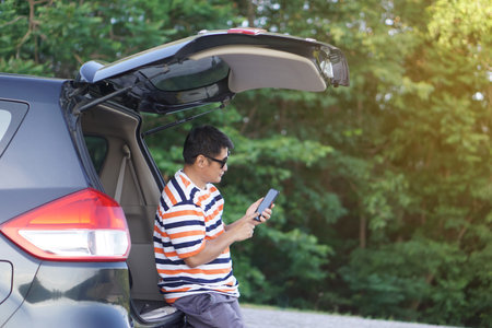 Happy Asian man traveler sits at the trunk of car, using smartphone. Concept, relax time. Vacation,...