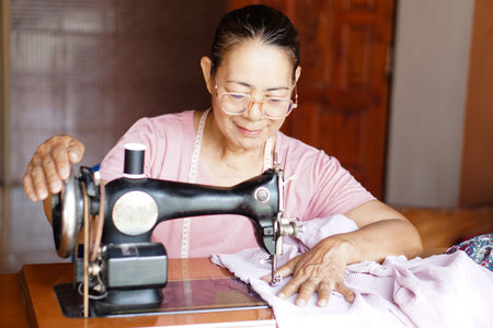 Happy senior Asian woman tailor is sewing cloth by vintage sewing machine at home. Concept, Senior  lifestyle, spend  free time to repair or design clothes. Hobby, not lonely.の写真素材