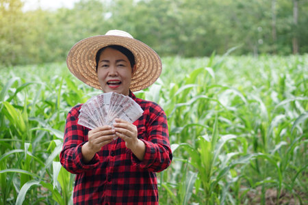 Happy Asian woman farmer is at garden, wears hat, red plaid shirt, holds Thai banknote money . Concept , Farmer happy to get profit, income, agriculture supporting money.の写真素材