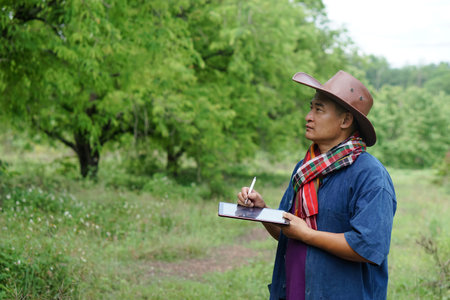 Asian male gardener is surveying and writing about growth and disease of tamarind plants on smart...