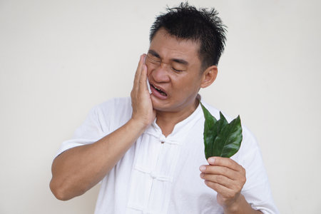 Asian man suffer from toothache, use hand to touch his cheek, close eyes, open mouth, holds herbal leaves. Concept, using natural herbs to remedy or relief pain symptoms.の写真素材
