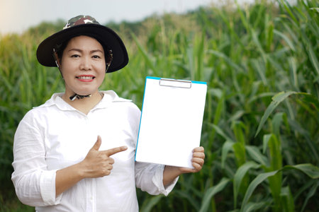 Asian woman farmer wears hat, white shirt, holds and point to blank paper clipboard, stands at maize garden. Concept. Agriculture occupation, survey and research to develop cropsの写真素材