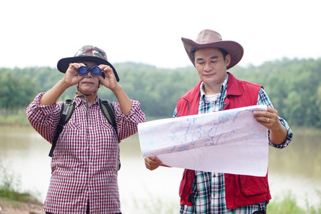 Asian man and woman travellers holds binoculars and paper map to explore natural route for trekking. Ecology study. Pastime activity, lifestyle. Explore environmentの写真素材