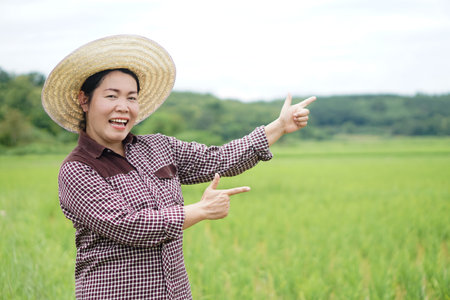 Happy Asian woman farmer is at paddy field, wears hat, plaid shirt, points fingers to present something. Concept, agriculture occupation, lifestyle. Copy space for adding textの写真素材