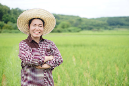 Asian woman farmer wears hat , plaid shirt, crossed arms on chest, stands at paddy field. Concept, agriculture occupation. Thai farmer lifestyle. Visit and take care of cropsの写真素材