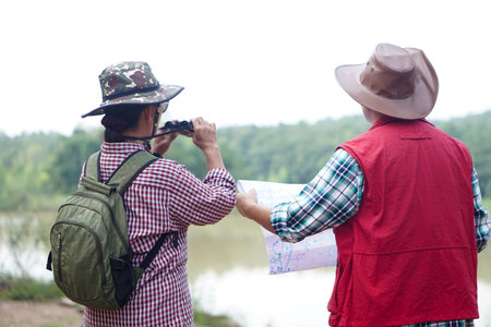 Back view of Asian woman and man travellers holds binocular and paper map to explore natural route...