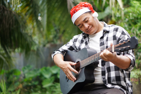 Asian man is playing acoustic guitar in the park, outdoor nature background. Concept, love music, hobby, recreation activity. Relax time. Playing music for celebrate Christmasの写真素材