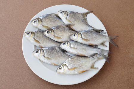 Raw fish for cooking on white plate, prepared for cooking, brown background. Concept, edible local freshwater fish from river in Thailand. Roach fish. Food ingredients.の写真素材