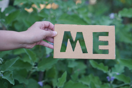 Close up hand hold brown paper cut to be word ME against green plants background. Concept, English teaching aid about object pronoun vocabulary lesson. Paper cutting calligraphy.の写真素材