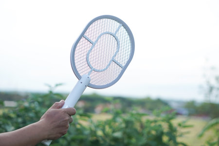 White electric mosquito swatter held firmly in hand, ready to zap away annoying insects in outdoor background. Modern, innovative device designed to eliminate mosquitoesの写真素材