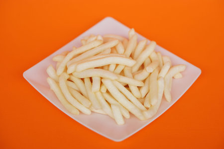 Crispy potato sticks on a white square plate, set against an orange background. Crunchy snack that...