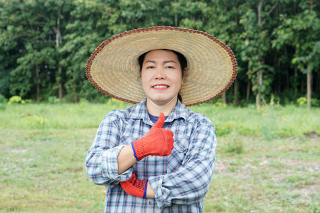 Asian female farmer wearing straw hat, plaid shirt, and red gloves smiling with thumbs up in green farmland. Confident rural woman representing agriculture, success, and sustainablの写真素材