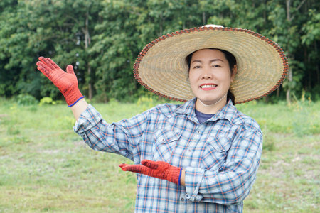 Smiling Asian female farmer wearing straw hat and gloves pointing and presenting with open hands in green field. Concept for agriculture, organic farming, rural lifestyleの写真素材