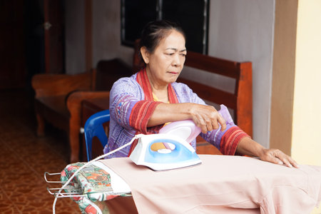 An elderly woman carefully irons clothing while spraying water to smooth the fabric. The warm, domestic scene reflects focus, routine, and the gentle rhythm of everyday home life.の写真素材