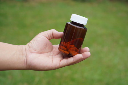 Hand holding brown glass bottle with capsules outdoors, representing herbal medicine, dietary...
