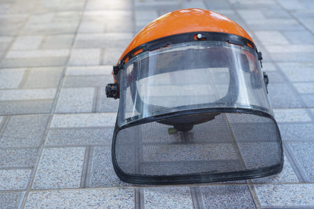 Orange safety helmet with full face shield placed on tiled floor outdoors, representing industrial protection, forestry and construction safety, professional protective equipment,の写真素材