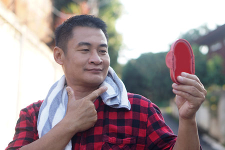 Middle aged man holding  small mirror and checking his face after work. Casual lifestyle moment with towel around neck, self care and personal grooming concept. Daily routineの写真素材