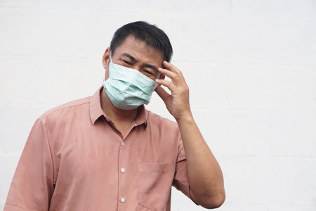 Asian man wears medical face mask touching his temple with tired expression, feeling headache, stress or illness. Concept of health problem, fatigue, virus protection, pandemicの写真素材