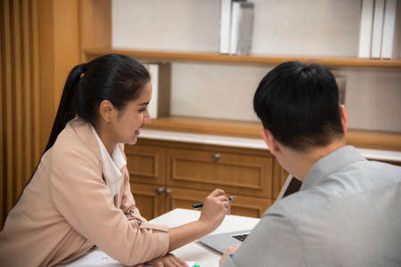 An Asian female executive and a business woman teaches a young male employee using a computer in their office. By photos from outside the officeの写真素材