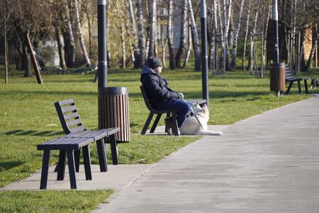 Krakow, Poland 01.12.2019: man in dark clothing sits on a bench in the Park on a bright Sunny autumn or spring day, a white dog lies on the ground at the feet of the owner. Family vacation with Pets.のeditorial素材