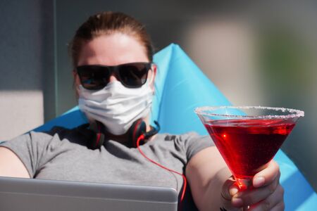 European girl with dark glasses and medical mask and cocktail works remotely or have virtual party on terrace or balcony on a Sunny warm day during quarantine or pandemic.Self-isolation,stay at home.の写真素材