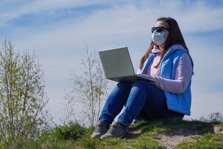 Young woman in medical mask works or studies remotely during pandemic on Sunny day sitting on grass...