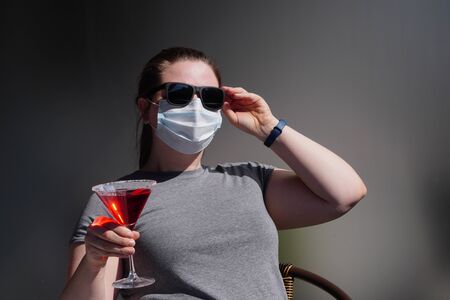 European girl with dark glasses and medical mask and cocktail works remotely or have virtual party on terrace or balcony on a Sunny warm day during quarantine or pandemic.Self-isolation,stay at home.の写真素材