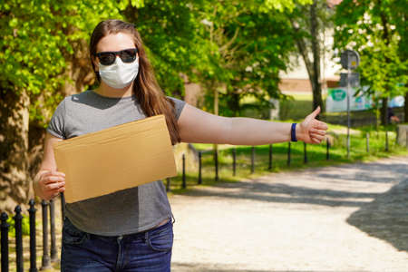 A girl in medical mask with empty cardboard sign stands by the road, catching car for Humorous hitchhiking trip. Getting out of quarantine, traveling after coronavirus. Pandemic fear.の写真素材