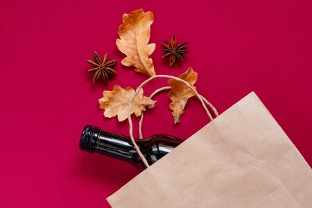 Paper bag with dark glass bottle of wine, alcohol present with dry leaves, composition. Hot vine aet for autumn. Flatly on maroon background, zero waste.Garbage recyclingの写真素材