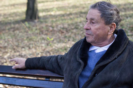 Portrait of a pensive senior man sitting on the bench, in the public park, outdoors. Old man relaxing outdoors and looking away. Portrait of elderly man enjoying retirementの写真素材
