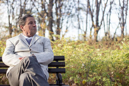 Portrait of a pensive senior man sitting on the bench, in the public park, outdoors. Old man relaxing outdoors and looking away. Portrait of elderly man enjoying retirementの写真素材