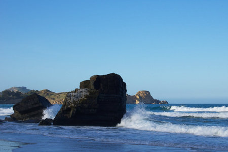Waves crashing against large basalt rocks on the beach around Seacliff-Karitane, New Zealand, sunlit rugged coastline in the backgroundの写真素材