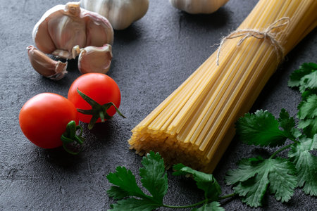 uncooked spaghetti pasta tied up with rope from burlap with cherry tomatoes, garlic heads and fresh branches of green cilantro on black backgroundの写真素材