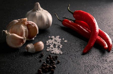 on a black background two heads and cloves of garlic opposite three red chili pepper between them track coarse sea salt and black peppercornsの写真素材