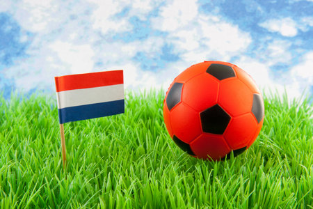 Orange ball and Dutch flag on soccer field against paper blue cloudy sky の写真素材