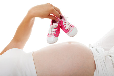 Woman is holding little pink shoes over pregnant belly on white background