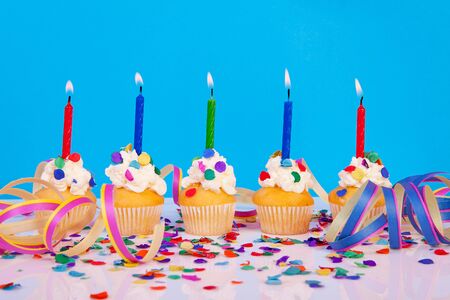 Birthday cupcakes with candles, party streamers and colorful confetti over blue backgroundの写真素材