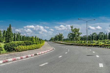 Beautiful asphalt road with green trimmed plants in the park on white cloud and blue sky background