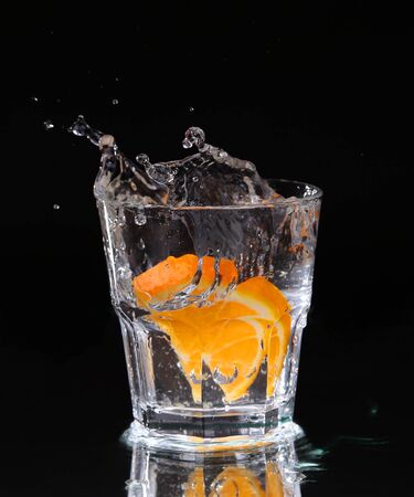 Slice of lemon splashing into a glass of water with a spray of water droplets in motion suspended in the air above the glass on a dark background.の写真素材