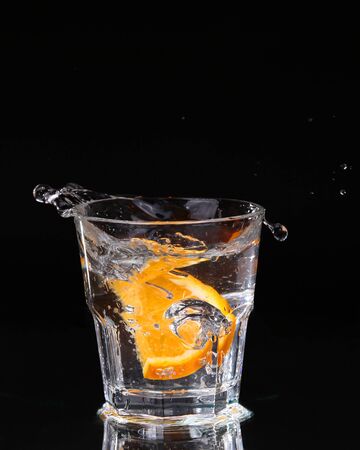 Slice of lemon splashing into a glass of water with a spray of water droplets in motion suspended in the air above the glass on a dark background.の写真素材