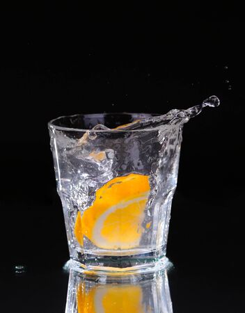Slice of lemon splashing into a glass of water with a spray of water droplets in motion suspended in the air above the glass on a dark background.の写真素材
