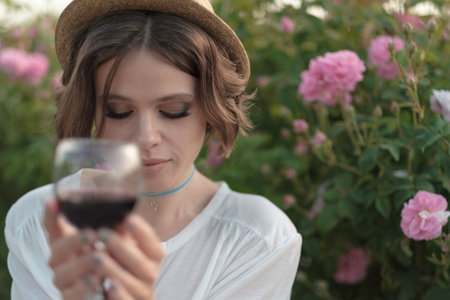 Beautiful young woman with curly hair posing near roses in a garden. Girl with a glass in her hand,...