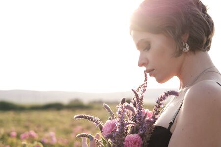 Beautiful young woman with curly hair posing near roses in a garden. The concept of perfume advertising.の写真素材